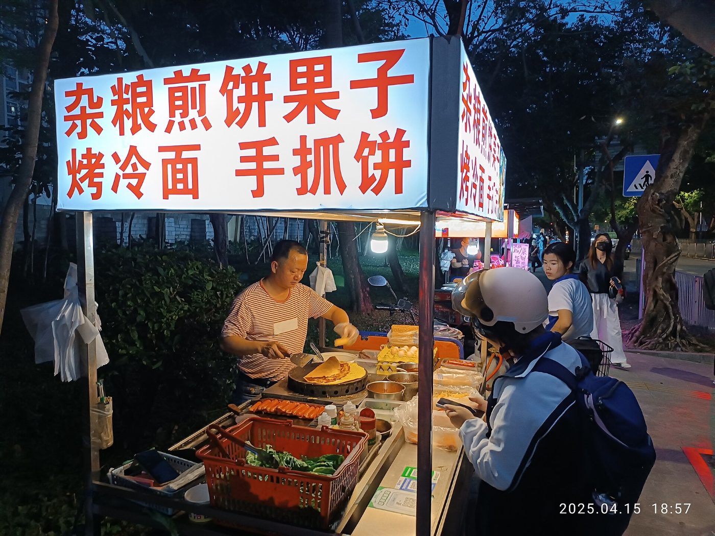 Wieczorny stragan z naleśnikami jianbing w Shenzhen: sprzedawca smaży naleśnik na okrągłej płycie, przed straganem czekają klienci
