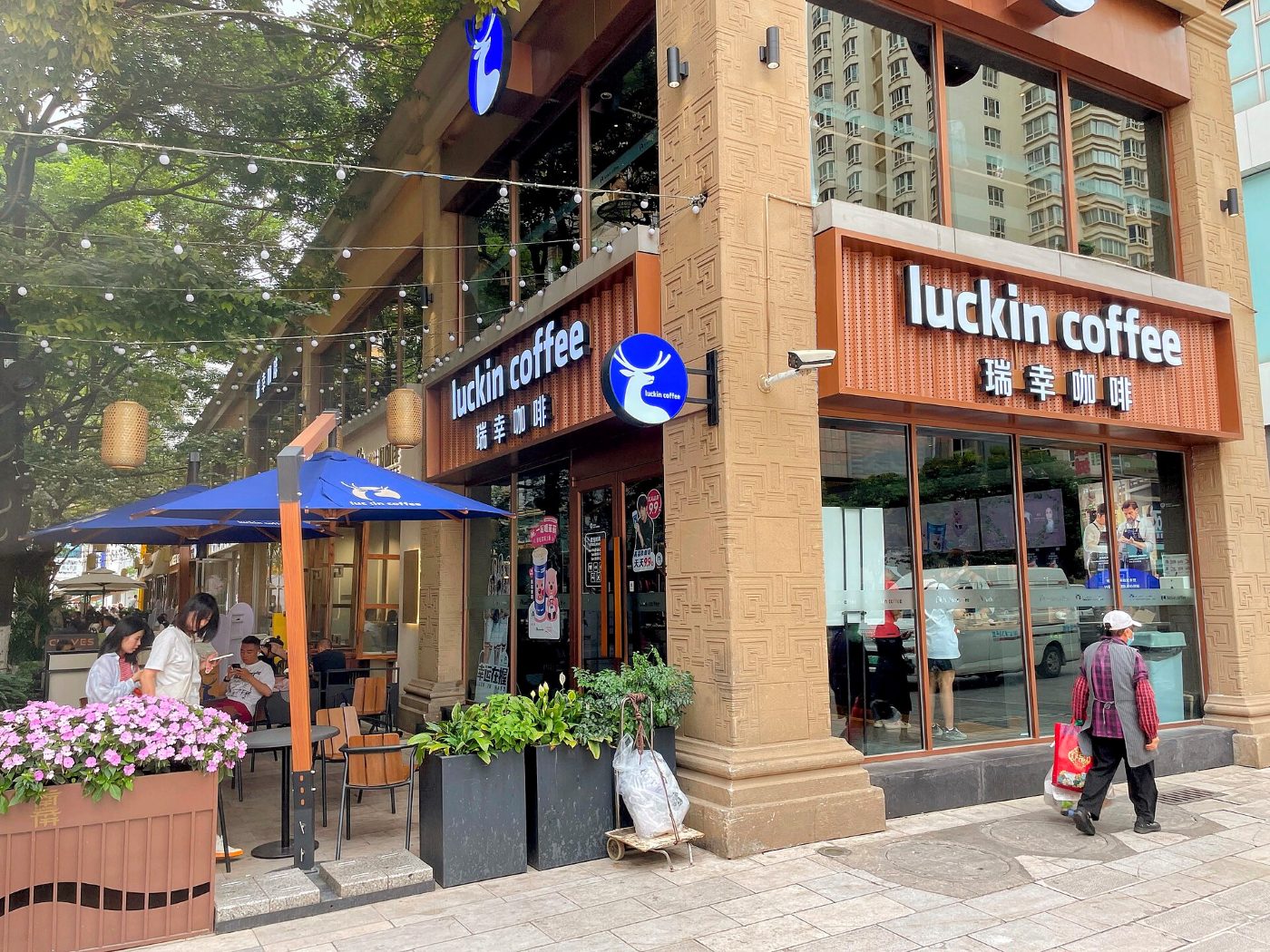 Kawiarnia Luckin Coffee w Kunmingu: dwie witryny z logo sieci, przed wejściem stoliki pod parasolami i kwiaty