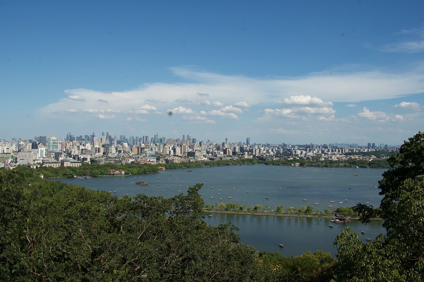 View from a hill over West Lake: the tree-lined Bai Causeway, boats on the water and Hangzhou's skyscrapers on the horizon