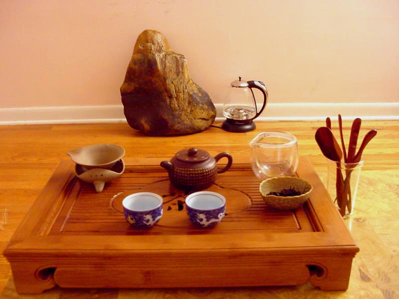 A wooden tea tray with drainage holding a clay teapot, two blue and white cups, a glass fairness pitcher, a strainer on a stand, a small basket of dry leaves and wooden tools in a cup