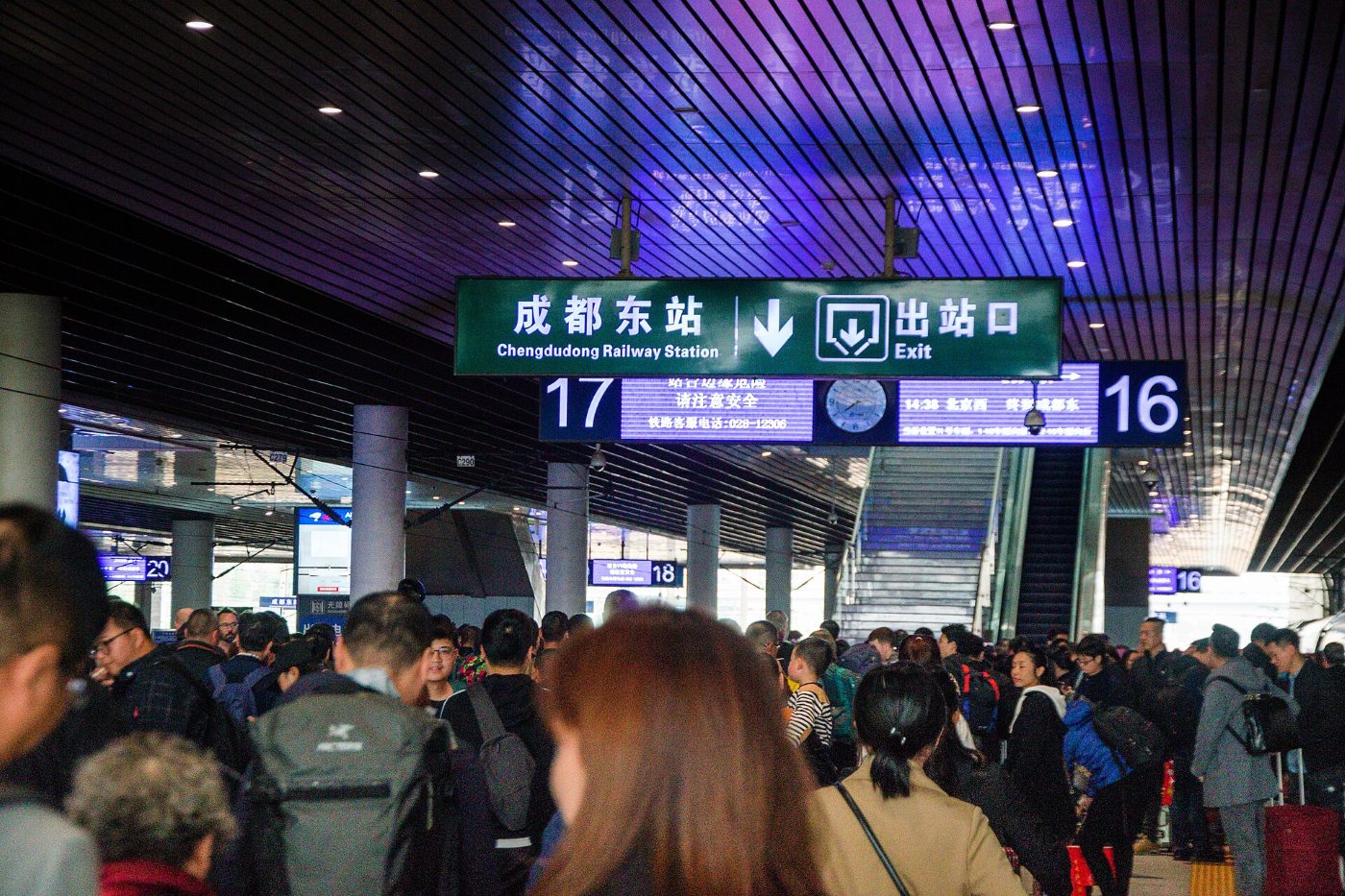 Podróżni w hali przyjazdów dworca Chengdu Dong pod dwujęzycznymi tablicami z nazwą dworca i numerami wyjść