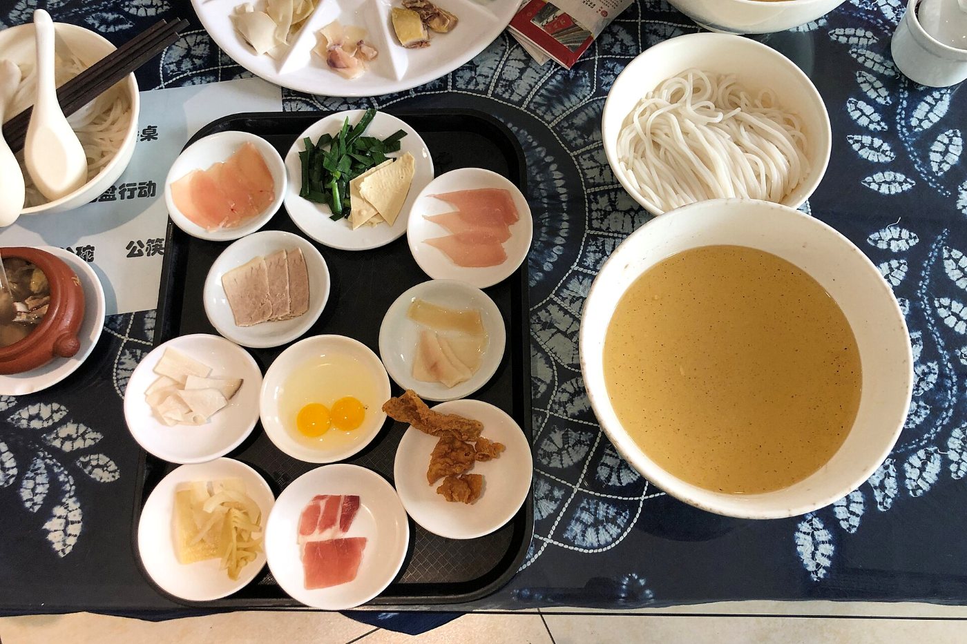A crossing-the-bridge noodle set: a bowl of broth, rice noodles and a dozen small dishes of raw toppings