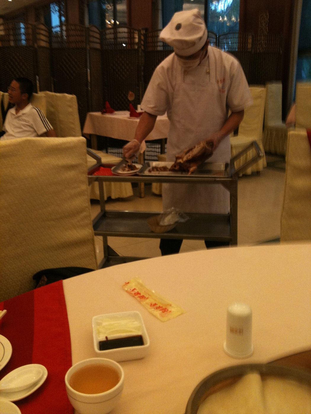 A chef in white uniform and cap carving a roast duck on a trolley beside a restaurant table