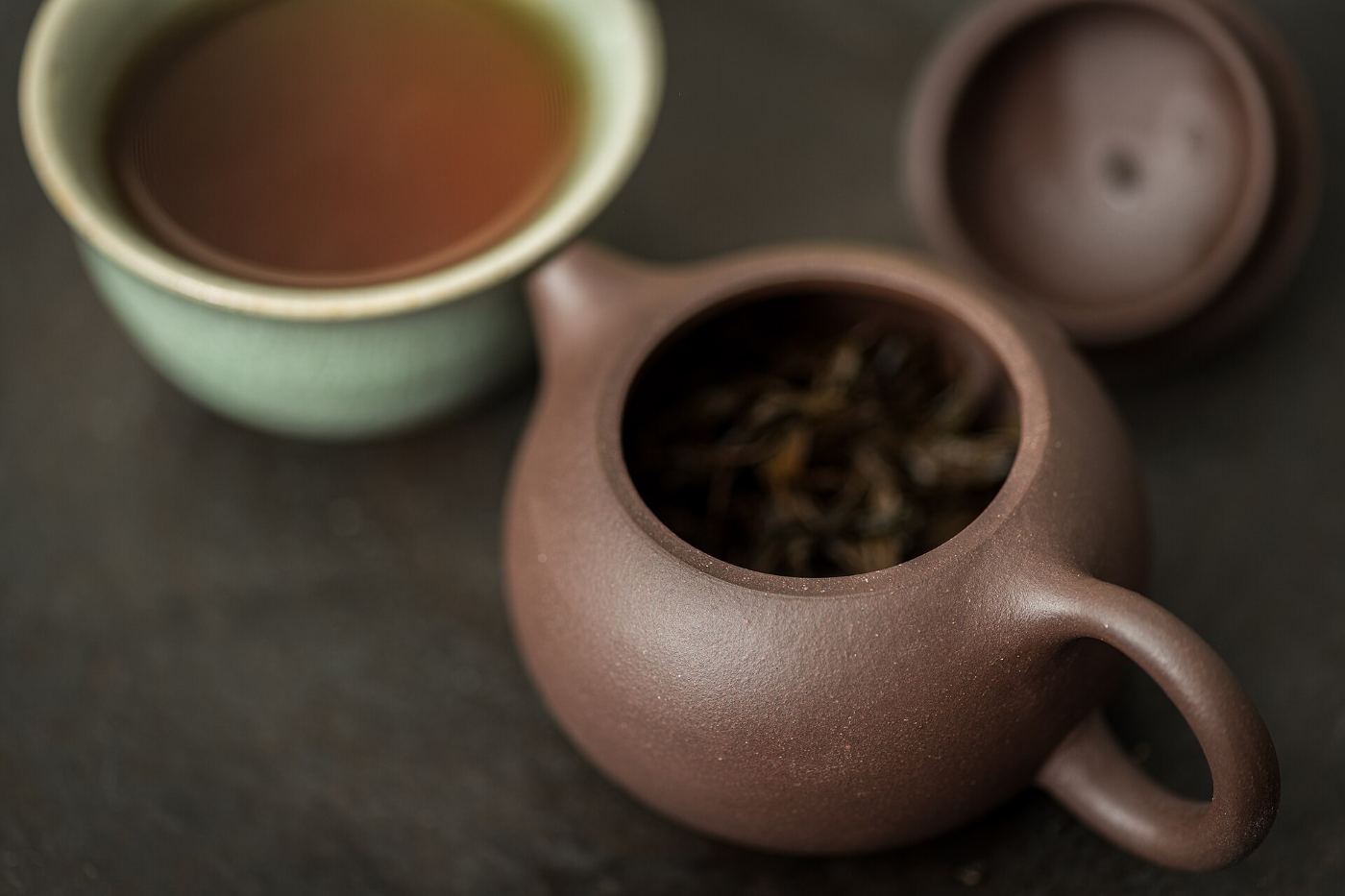 A brown Yixing clay teapot with black tea leaves inside and a green cup of tea