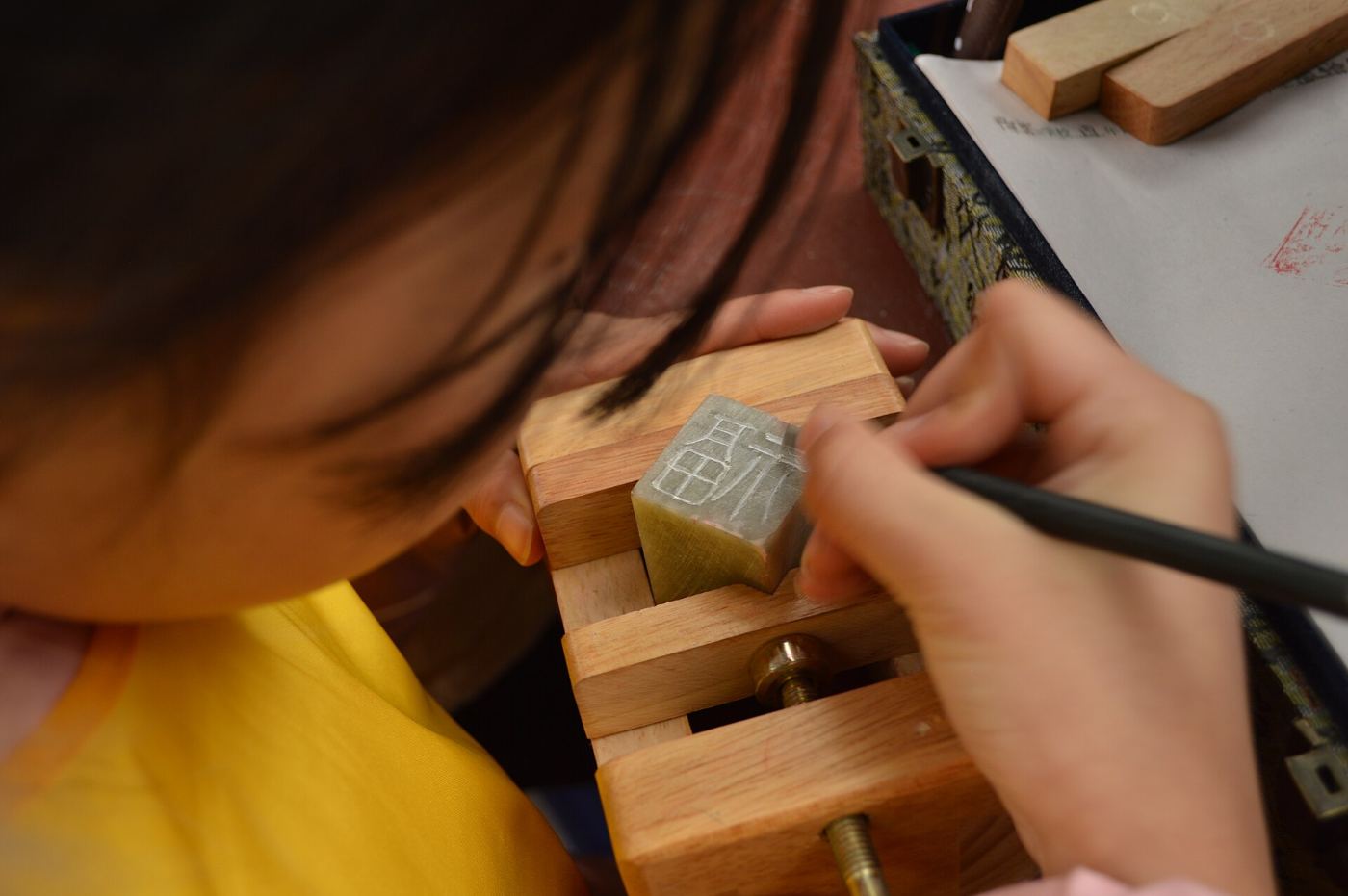 A craftsman carving Chinese characters into a stone seal held in a small wooden vice