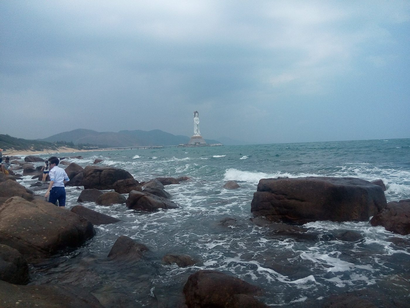 A rocky shore with waves and, far out at sea, the white Guanyin statue at Nanshan