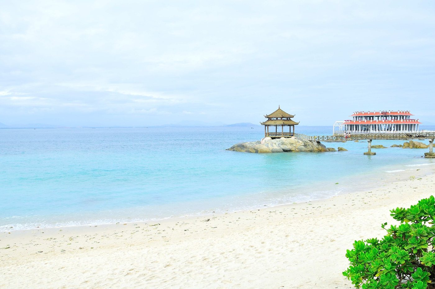 A white beach and turquoise water on Wuzhizhou Island, with a pavilion on the rocks and a pier behind