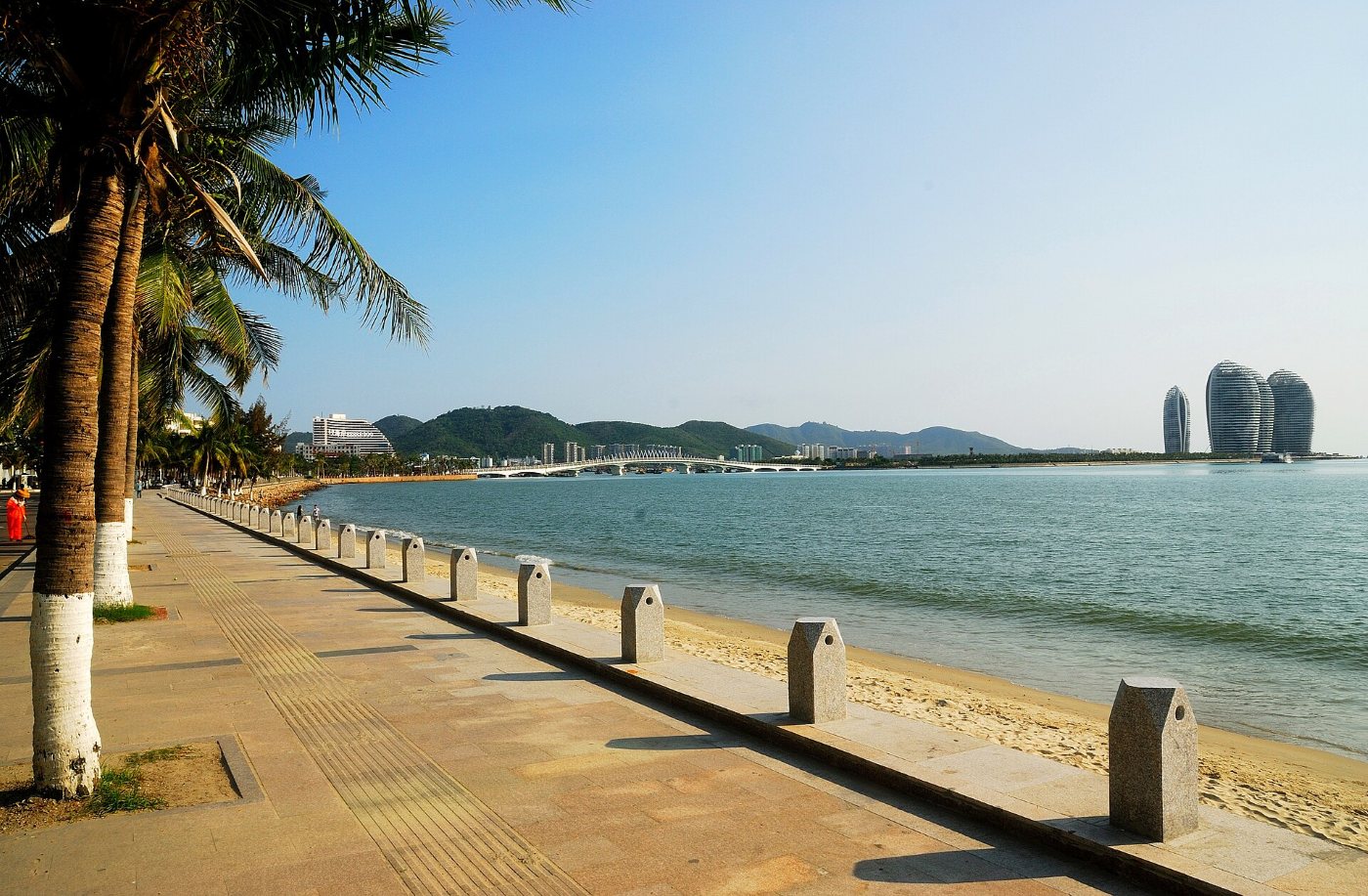 Seaside promenade lined with palm trees on Sanya Bay, with a bridge and the oval towers of Phoenix Island behind