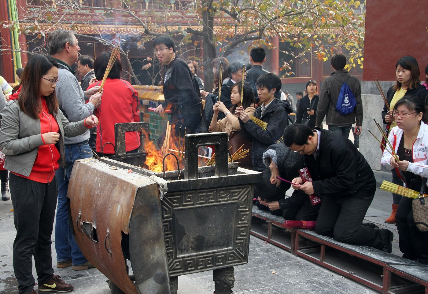 People holding bundles of lit incense sticks by a large incense burner in a courtyard of the Lama Temple, some kneeling on a wooden platform