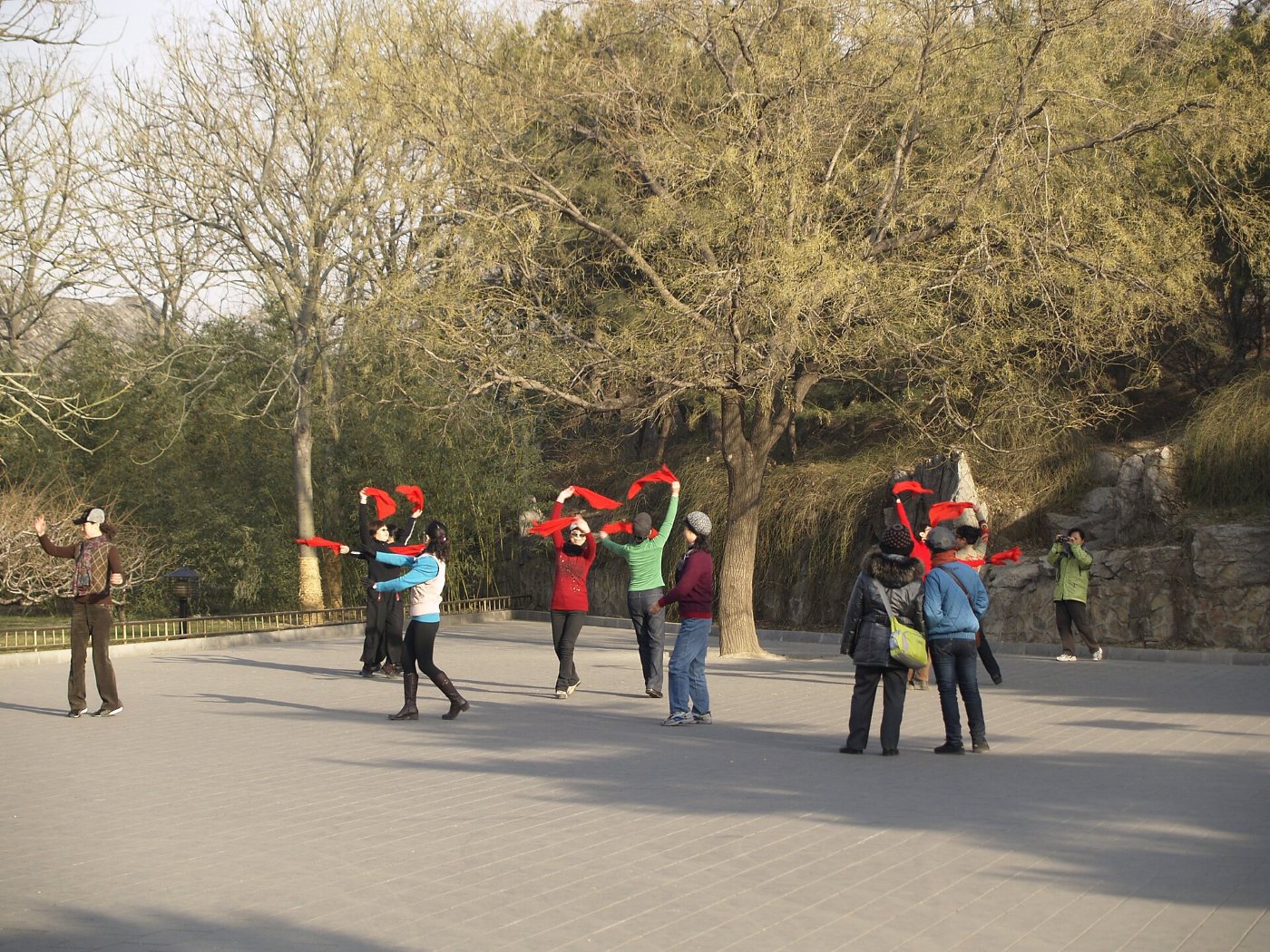 A group of women dancing with red scarves on a square in a park, with passers-by and a photographer nearby