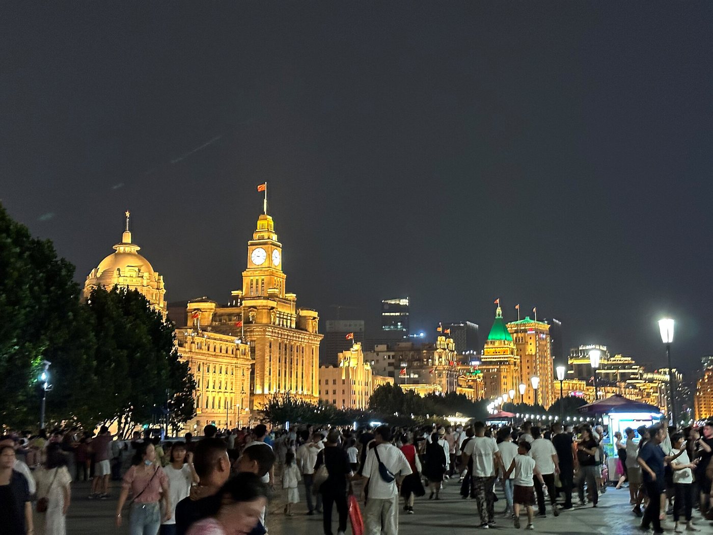 The illuminated colonial buildings of the Bund with a clock tower, and crowds on the promenade