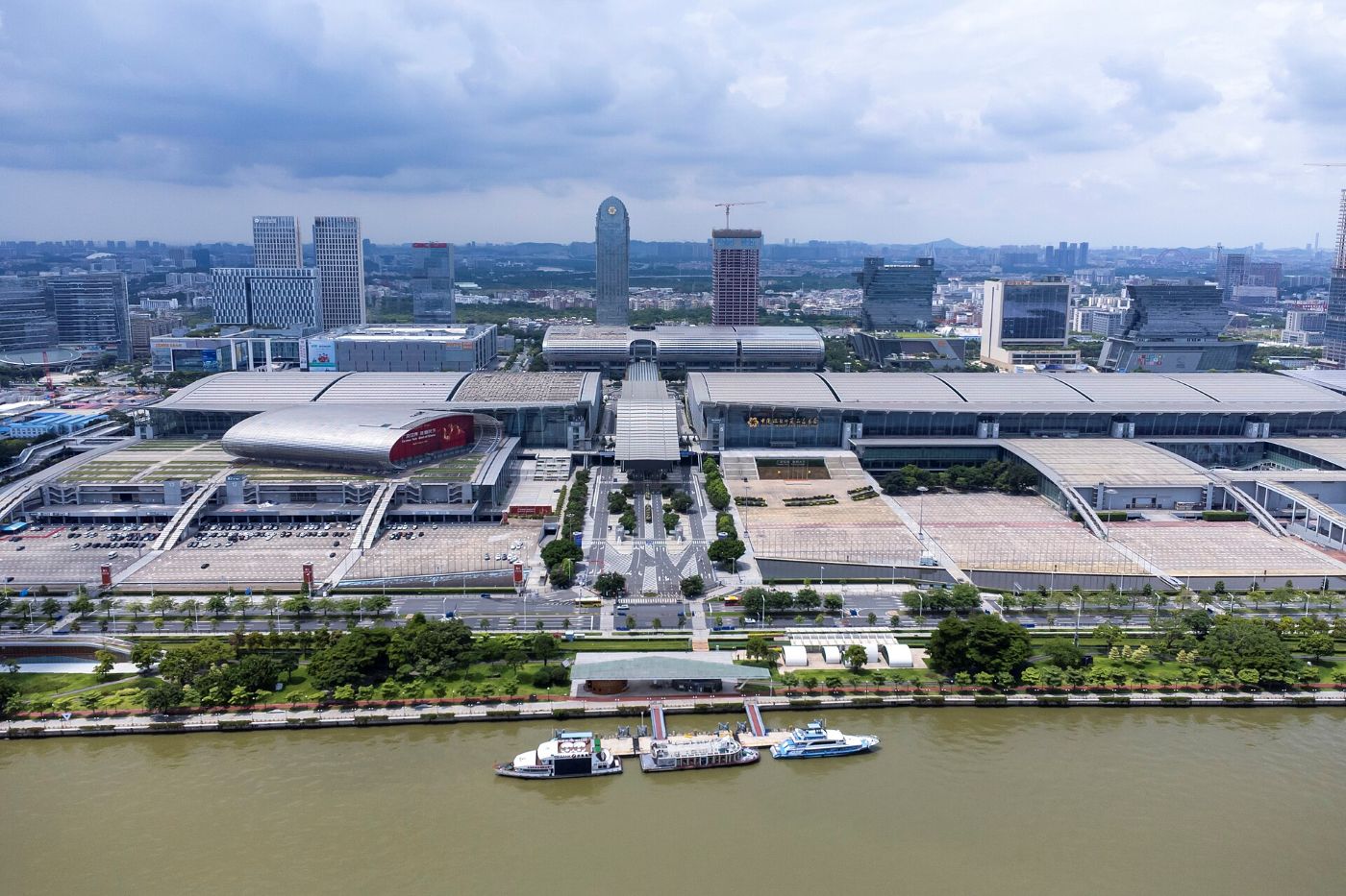 The Pazhou exhibition complex from the air: several halls by the Pearl River, a pier with ferries in front and skyscrapers behind