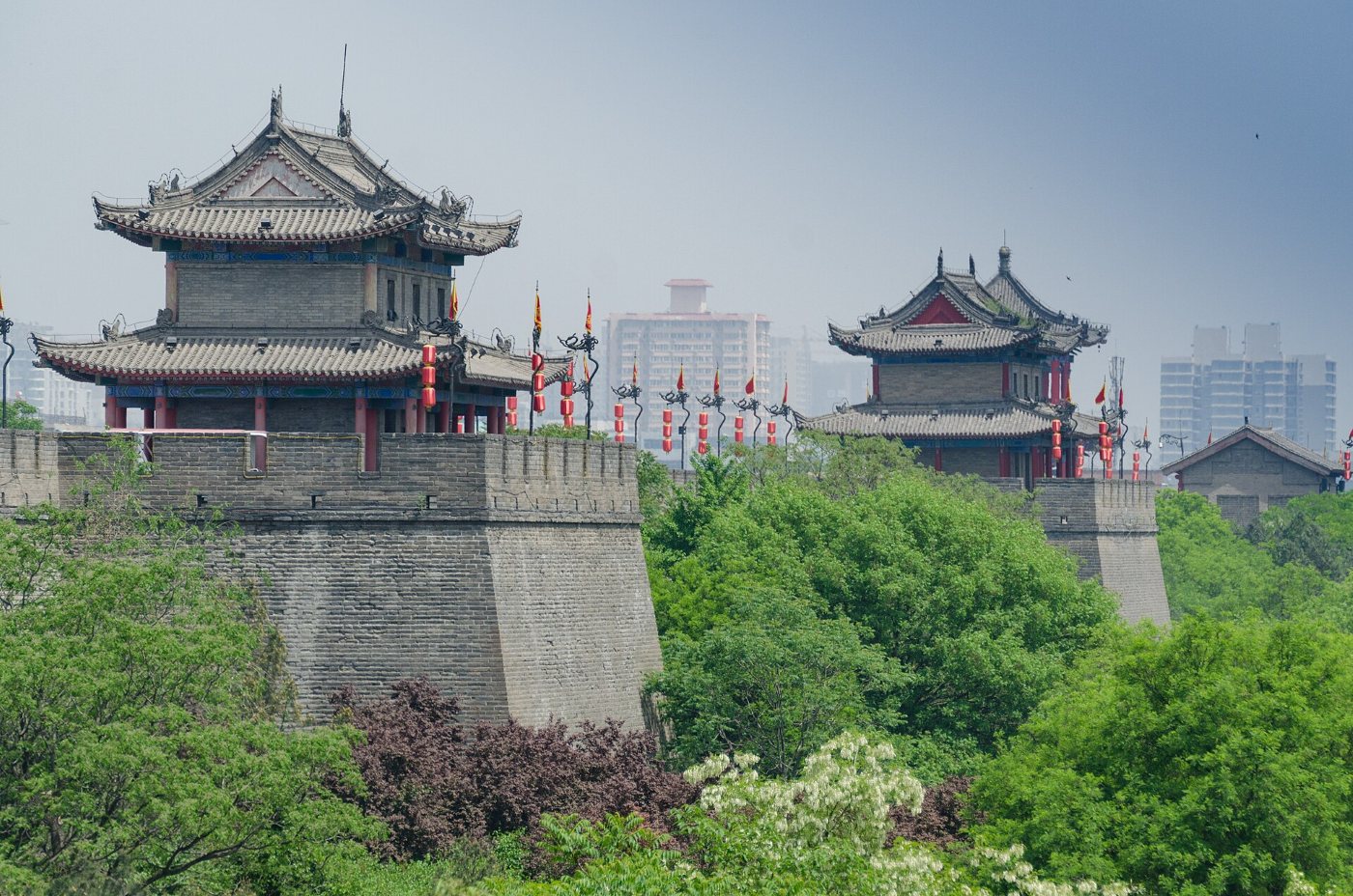 Xi'an city wall with two watchtowers with curved roofs, red lanterns and flags, green trees below