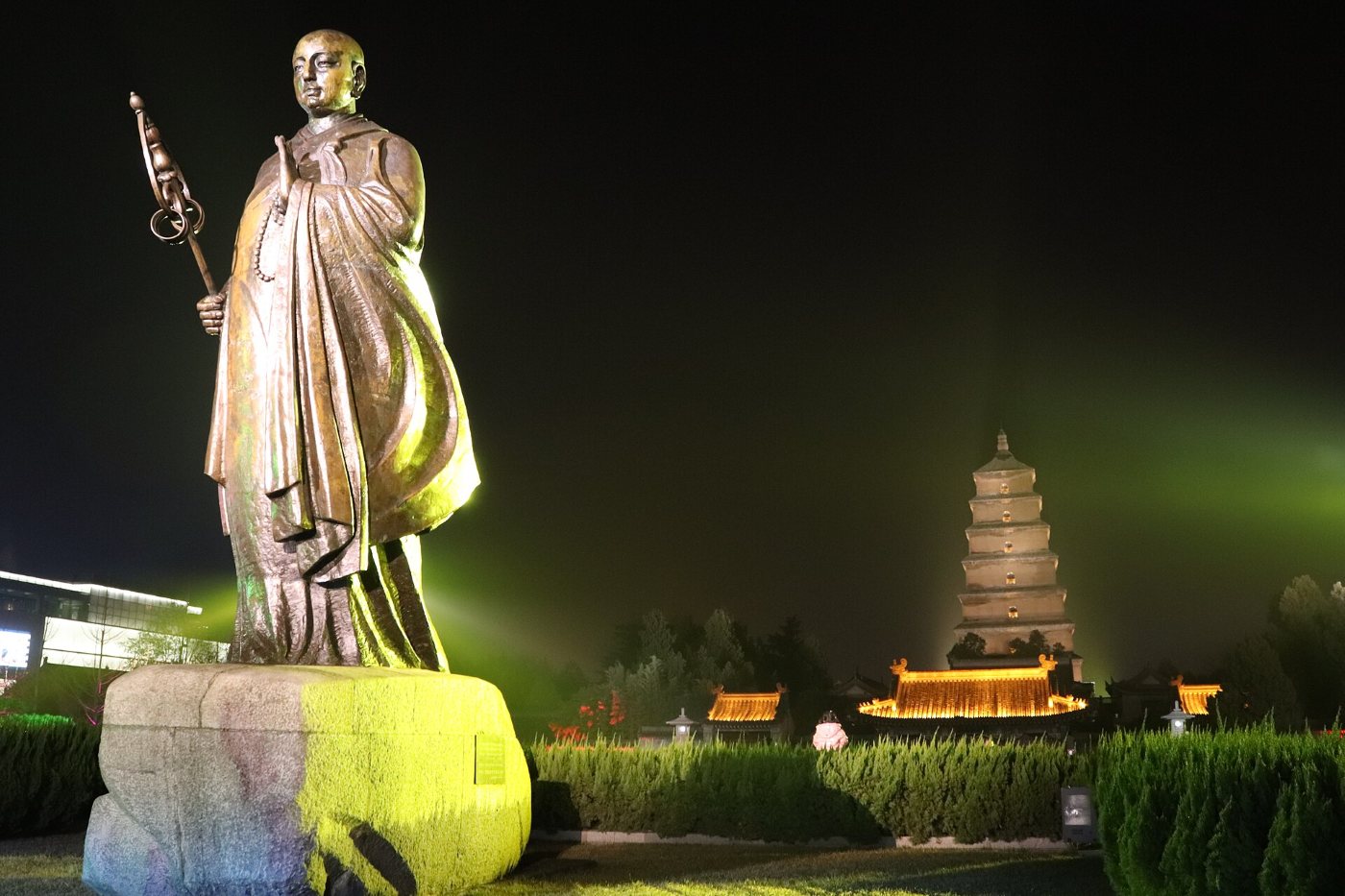 A statue of the monk Xuanzang on a stone plinth in the south square, the illuminated Big Wild Goose Pagoda behind at night