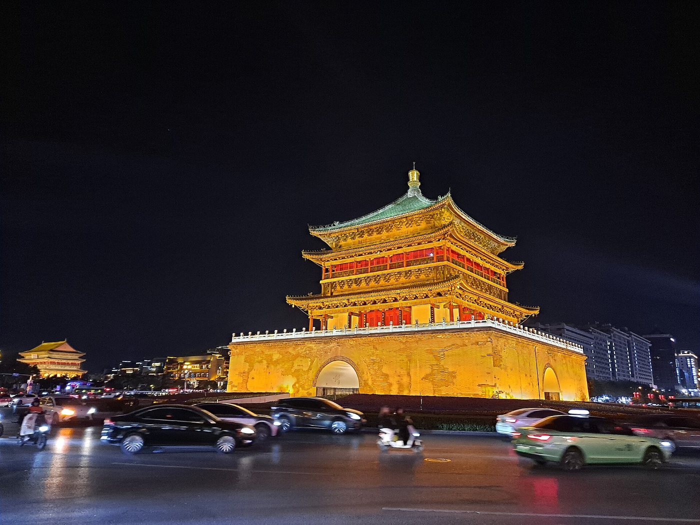 The Bell Tower in Xi'an at night: a golden-lit tower with a green roof on a stone base, cars on the roundabout in front, the Drum Tower on the left