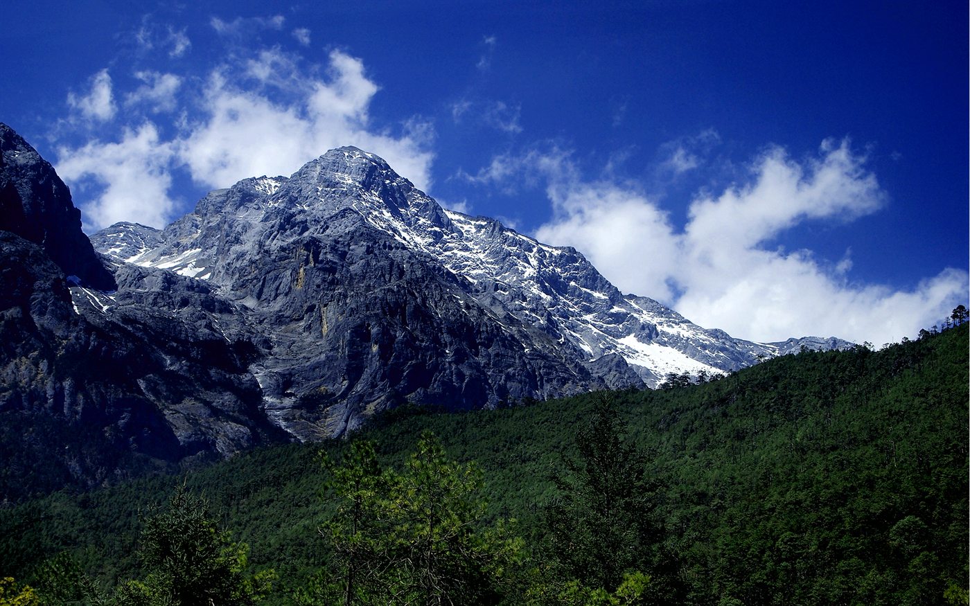 The snowy, rocky summit of Jade Dragon Snow Mountain above a forested slope, against a blue sky