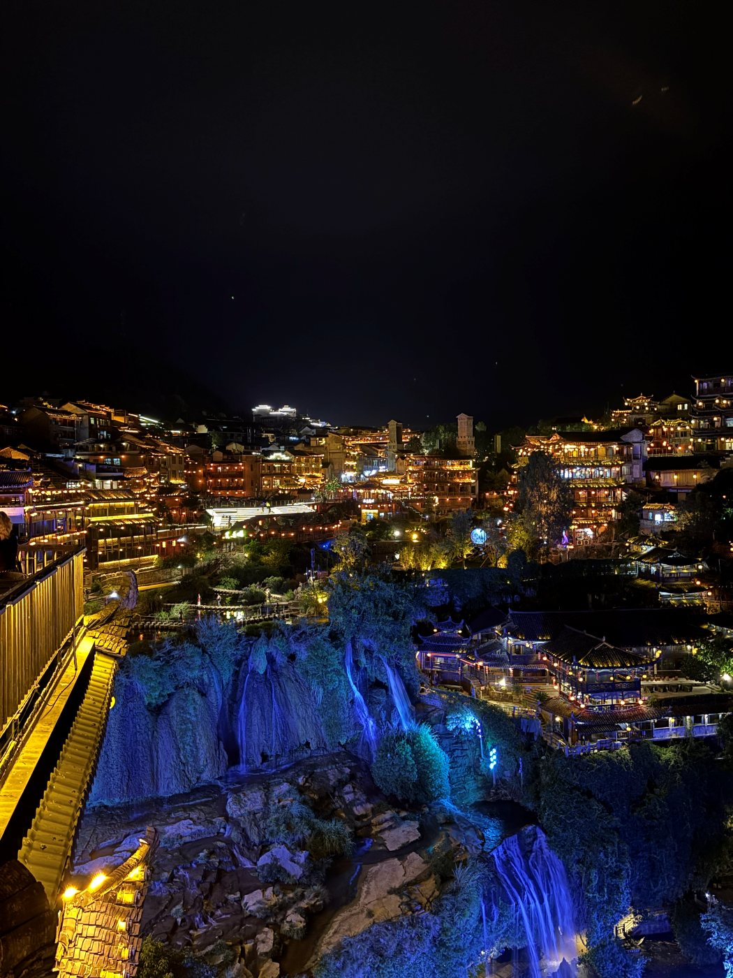 Furong at night: a blue-lit waterfall flowing beneath illuminated houses on the edge of a cliff