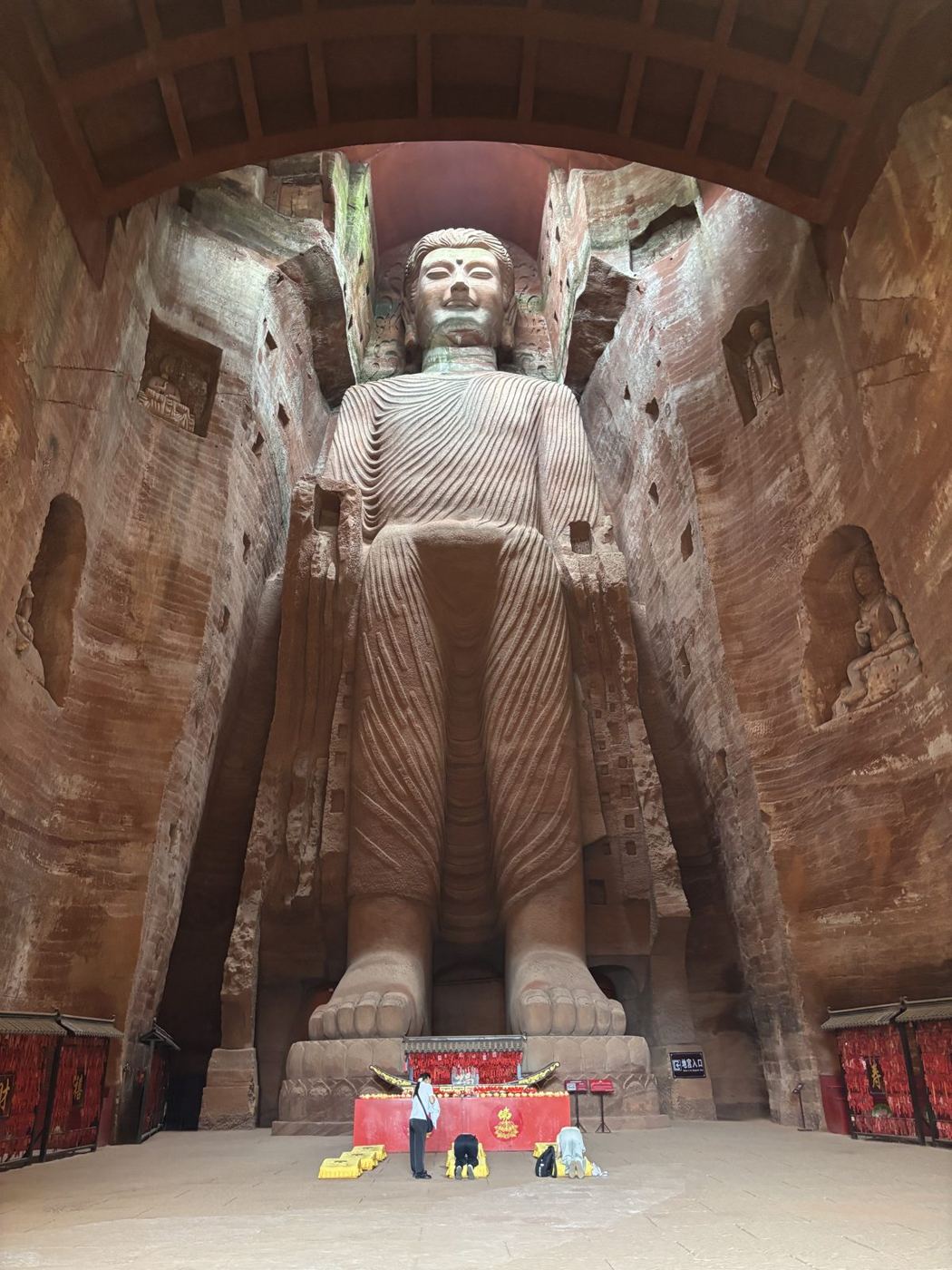 A tall standing Buddha carved into a rock niche, with an altar and kneeling worshippers at its feet