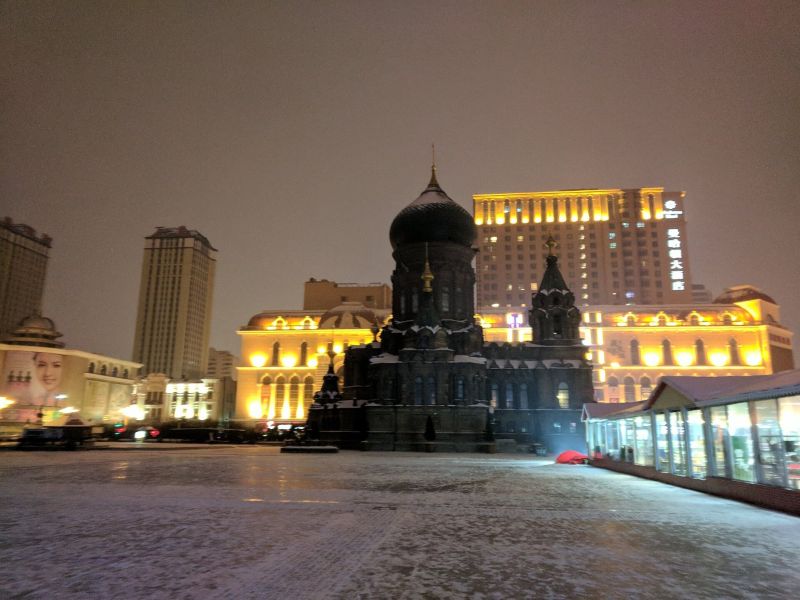 Sobór św. Zofii w Harbinie zimą nocą: ciemna kopuła na tle podświetlonych kamienic, plac pokryty śniegiem