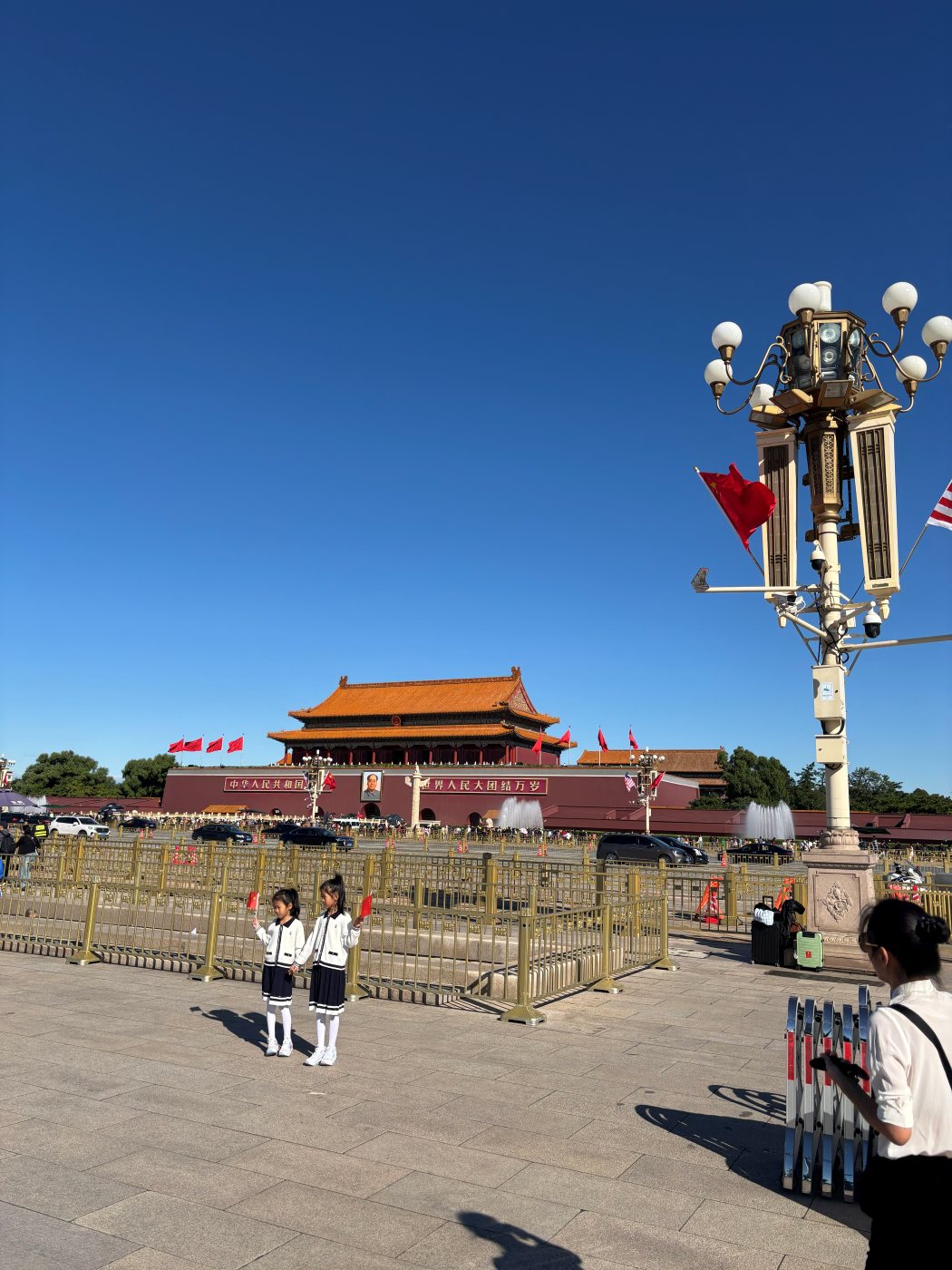 The Tiananmen Gate with Mao's portrait seen from the square, a lamp post with flags and visitors in the sunshine in the foreground