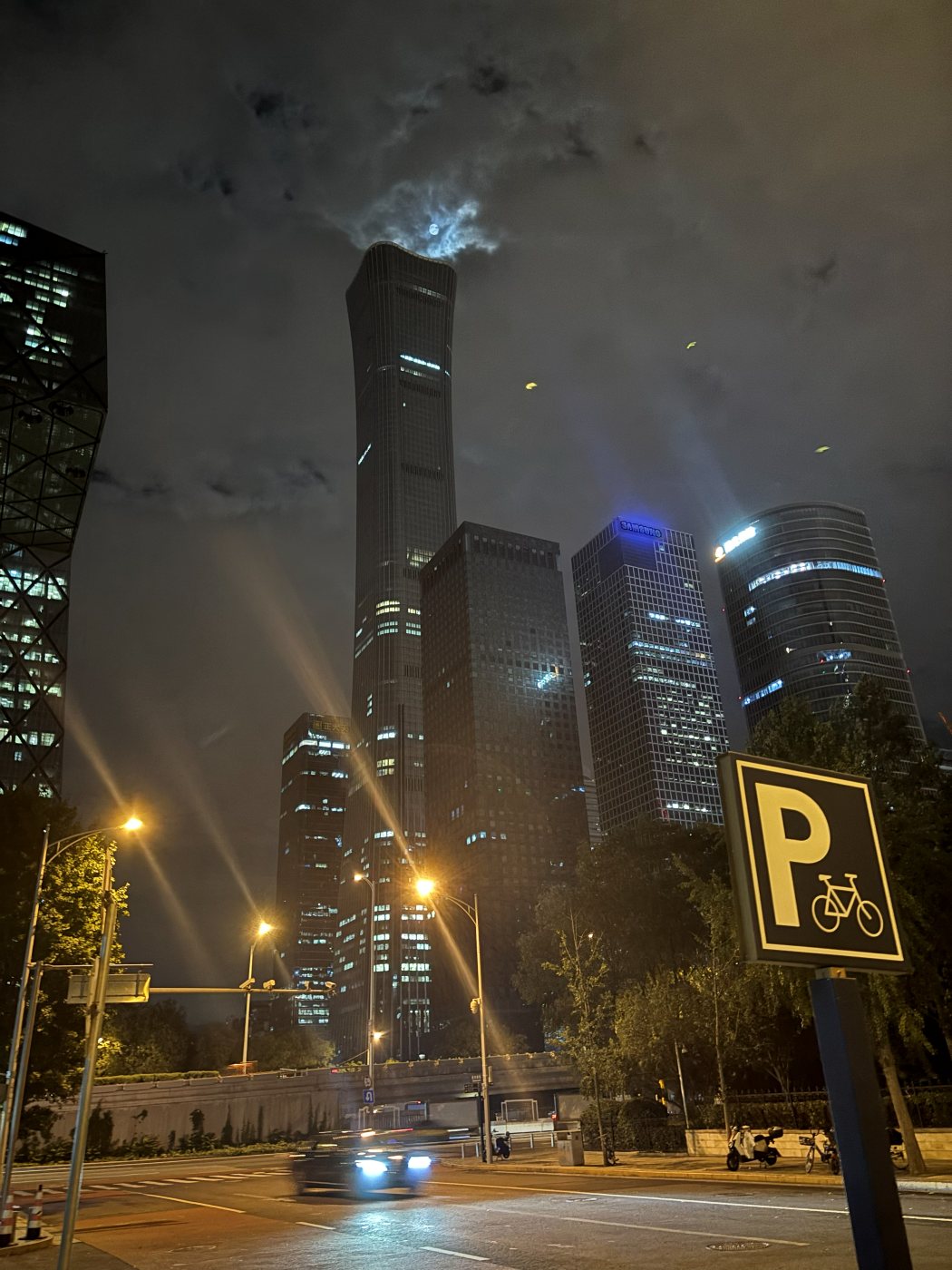 Skyscrapers of Beijing's CBD at night, a street and a bicycle parking sign in the foreground
