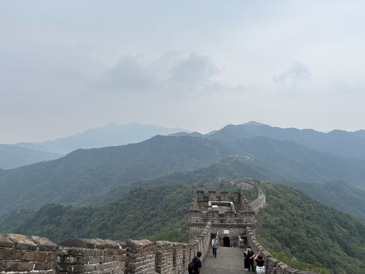 The Great Wall with a stone watchtower running across forested mountains near Beijing, with tourists on the wall