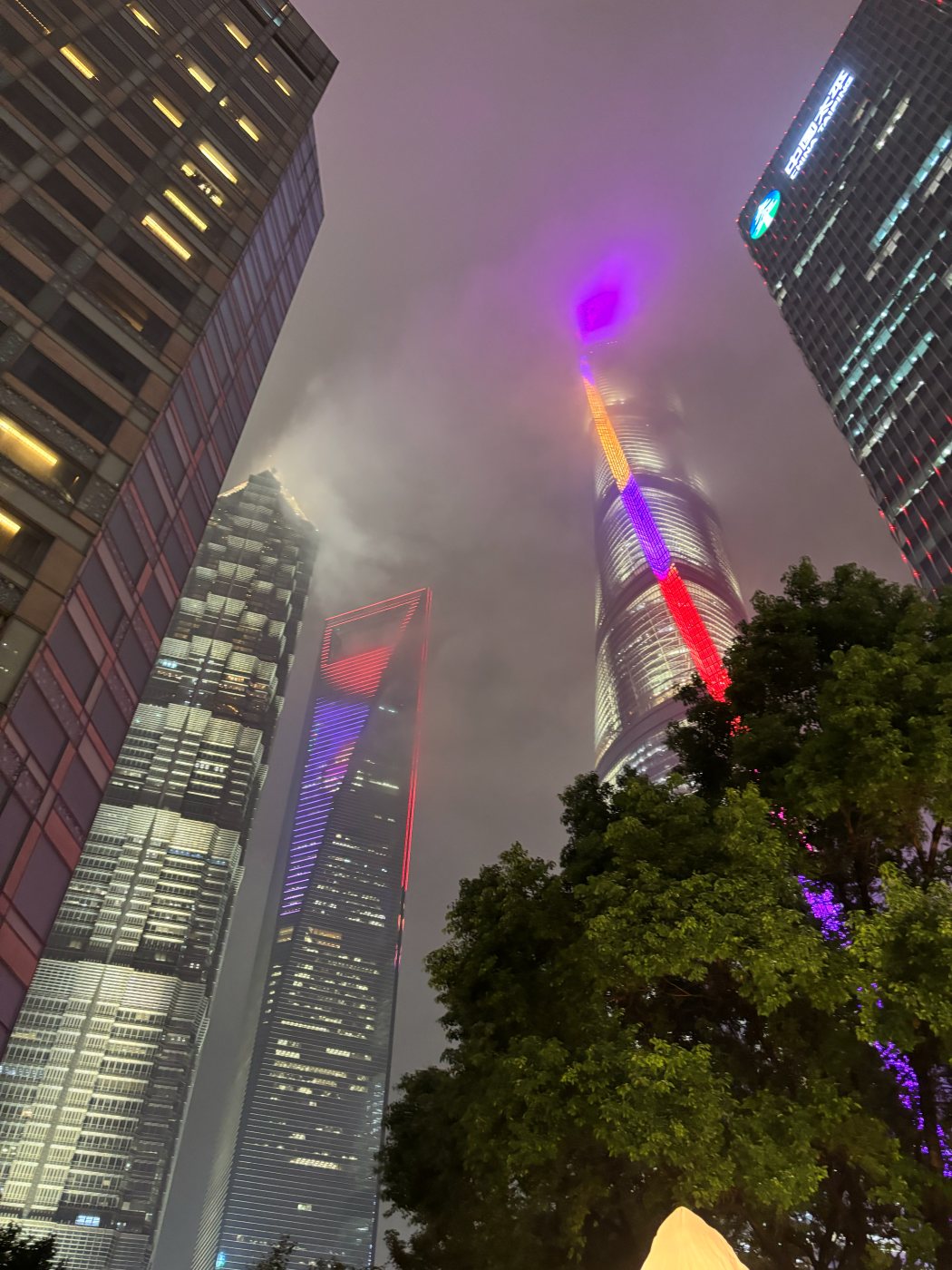 Lujiazui skyscrapers at night seen from below, their tops lit in colour against the clouds