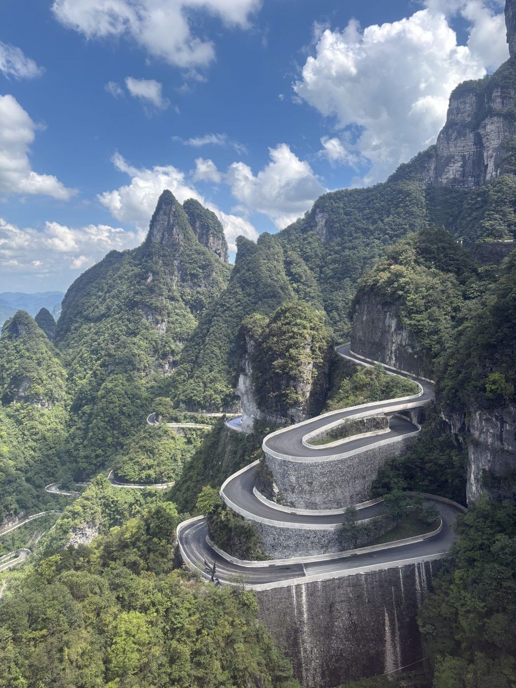 The 99-bend road on Tianmen Mountain: a road winding up a green slope between rocks, clouds above
