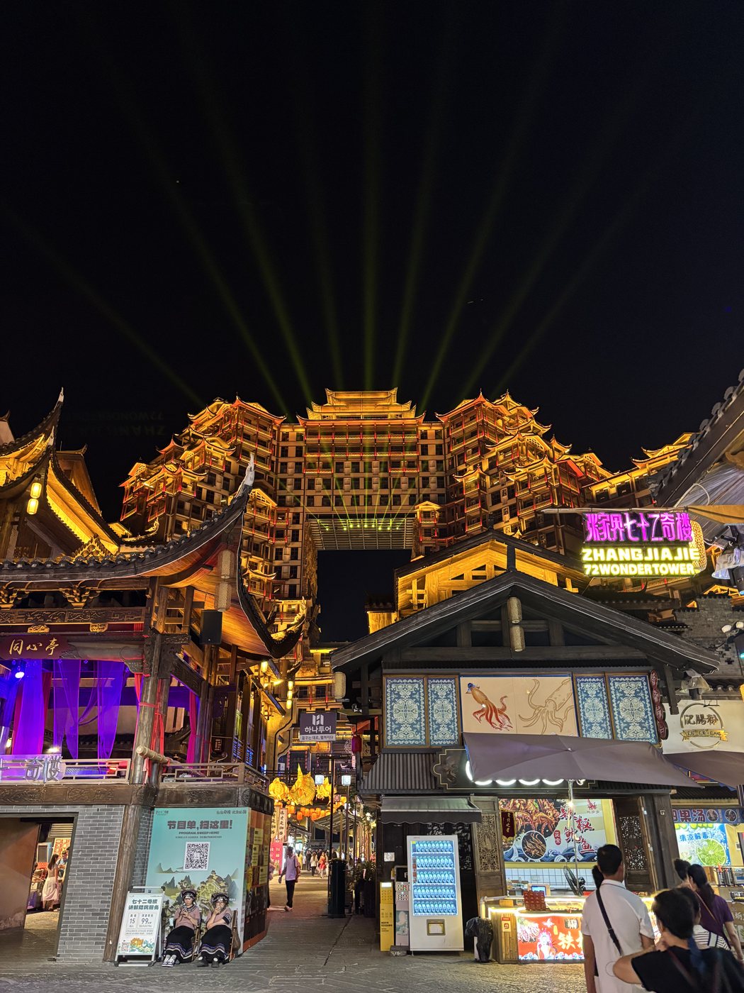 The illuminated 72 Qilou complex in Zhangjiajie at night, with a shopping street and a QR code poster in front
